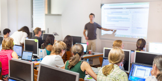 Viele Menschen sitzen in einem Kursraum vor Computern. Vorne steht ein Lehrer und zeigt etwas auf einem Bildschirm.