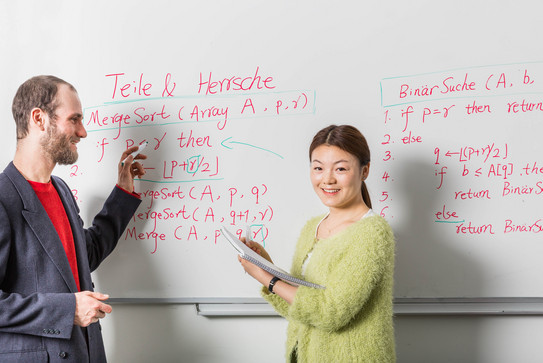 Lehrsituation-Whiteboard Zwei Personen stehen vor einem Whiteboard mit Formeln und Beschriftung. Eine Person hat einen Stift in der Hand, die andere einen Block.
