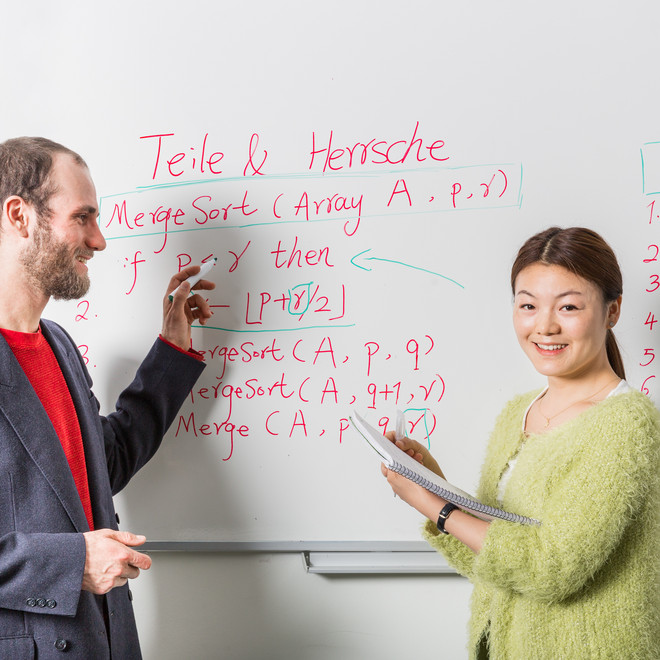 Zwei Personen stehen vor einem Whiteboard mit Formeln und Beschriftung. Eine Person hat einen Stift in der Hand, die andere einen Block.